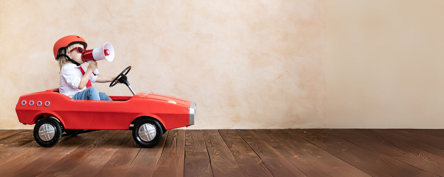 Kind rijdt in een rode speelgoedauto en roept vrolijk door een megafoon — symbool voor de Sproutz Sale vol stijlvol en duurzaam speelgoed.