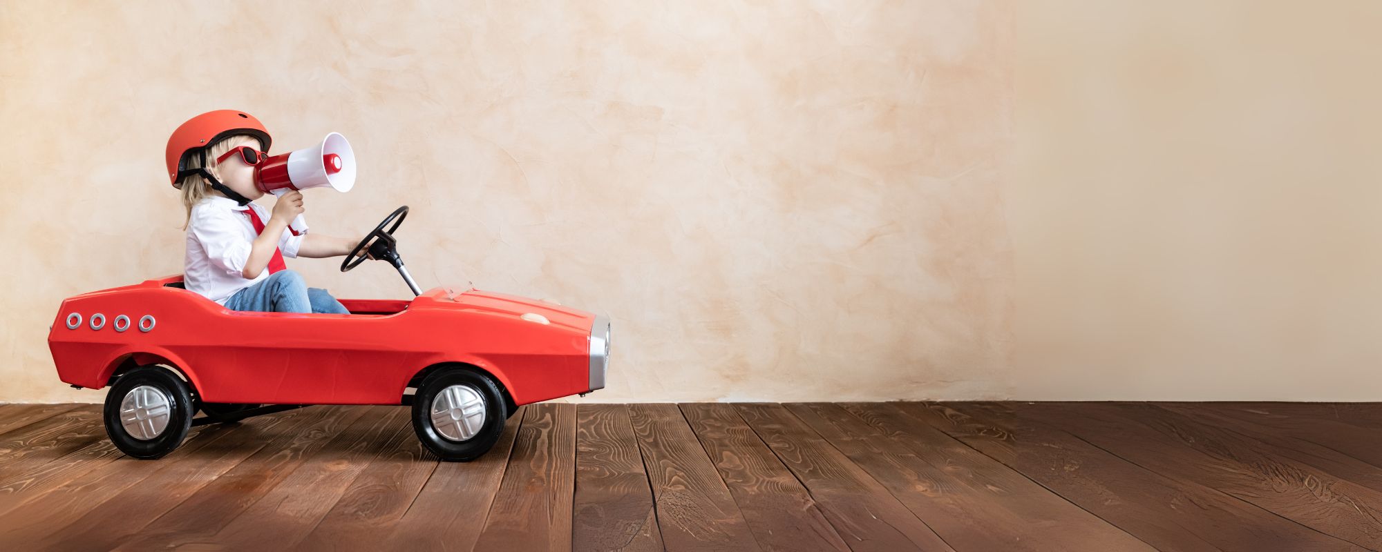 Kind rijdt in een rode speelgoedauto en roept vrolijk door een megafoon — symbool voor de Sproutz Sale vol stijlvol en duurzaam speelgoed.