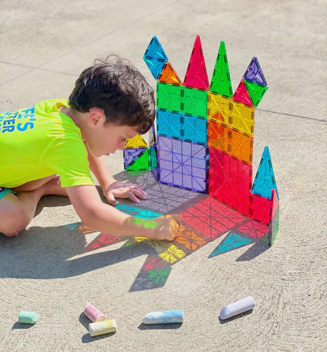 Kind speelt met een hondfiguur en bouwt een kleurrijk huis van magnetische Magna Tiles op een poef in een gezellige woonkamer.