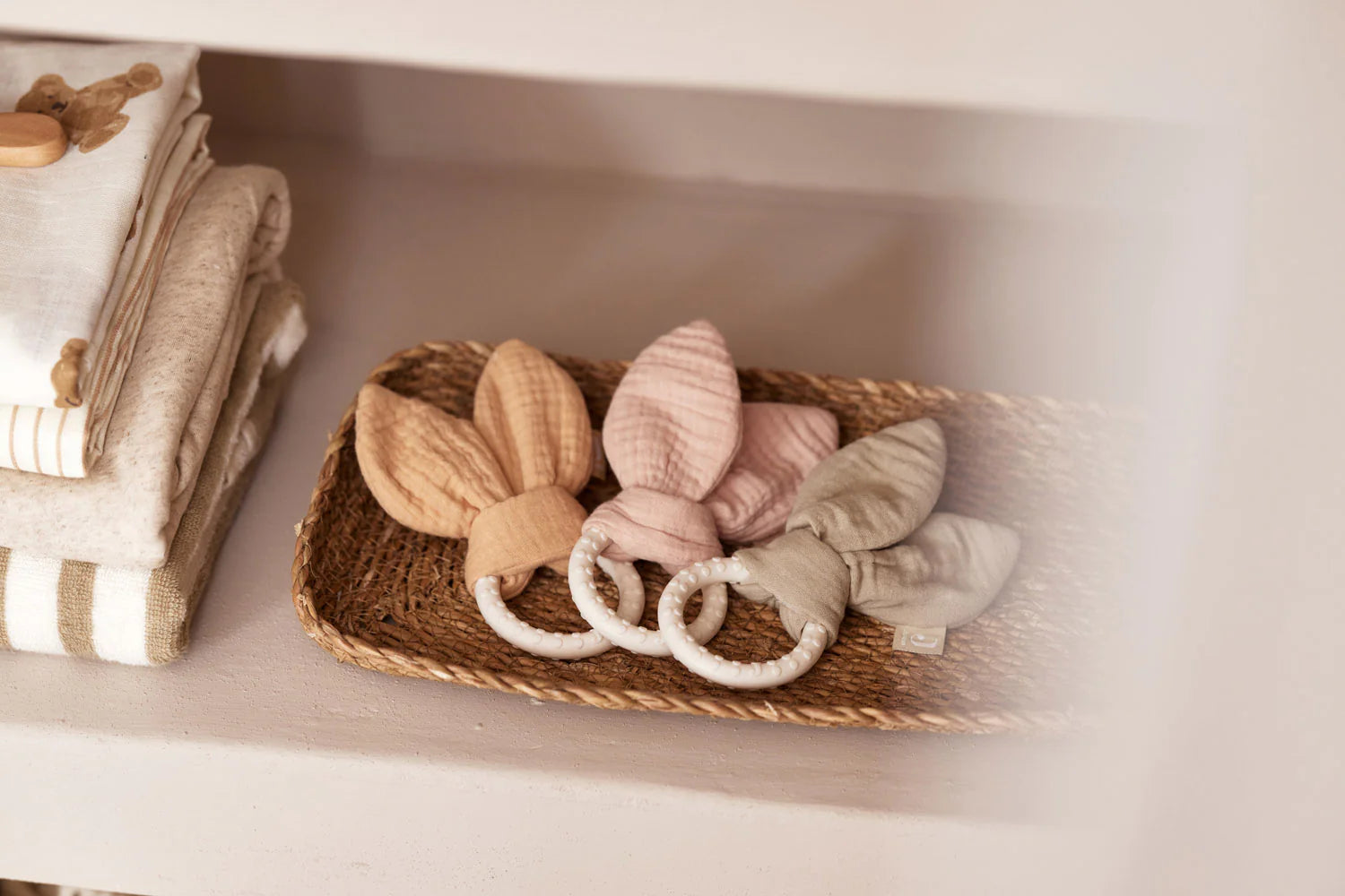 Jollein Bijtring Siliconen Bunny Ears in drie kleuren in een rotan mandje naast opgevouwen babytextiel, baby cadeau.