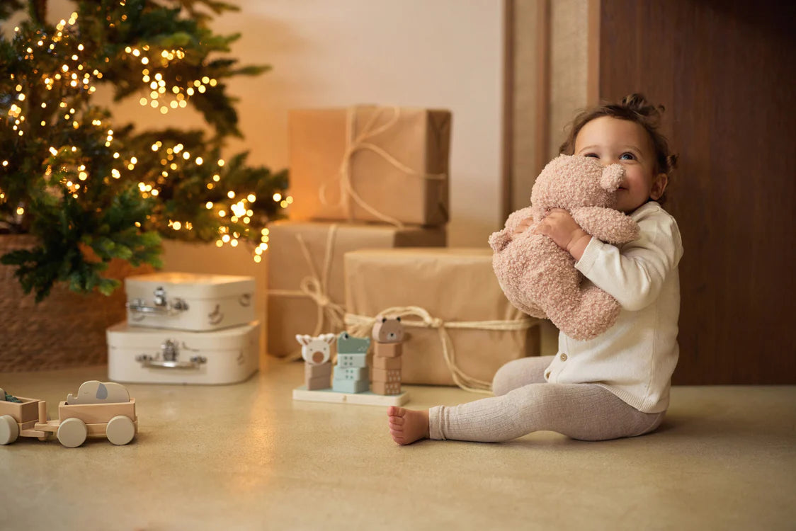 Baby knuffelt een Jollein teddybeer in wild rose bij de kerstboom, zachte dikke knuffel als warm cadeau.