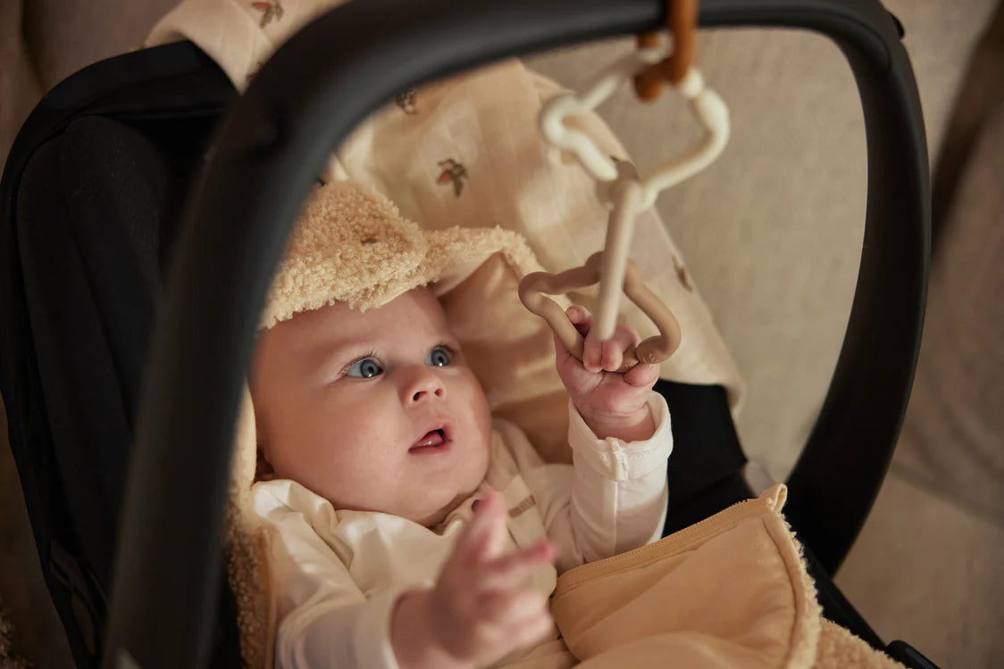 Baby reikt naar opgehangen Jollein Teddy Bear bijtringen in autostoel, ideaal kraamcadeau en sensorisch baby speelgoed.