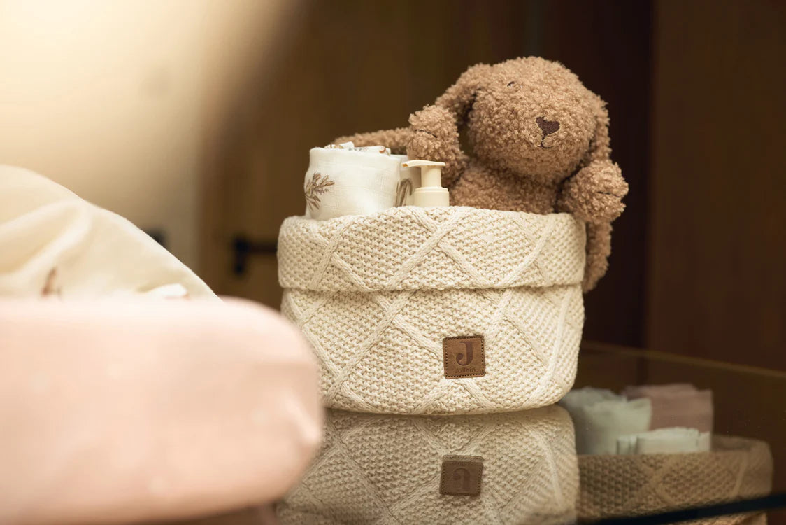 Biscuitkleurige Jollein Knuffel Bunny in gebreid mandje met babyverzorgingsproducten in warme sfeer.