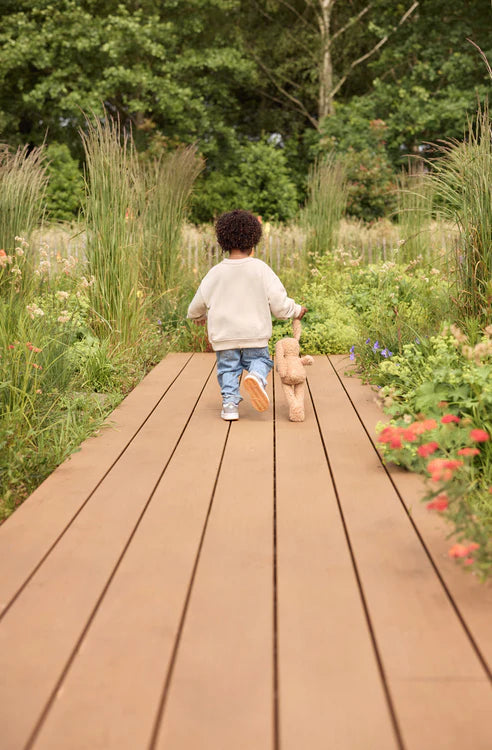 Kind loopt met biscuitkleurige Jollein Knuffel Bunny over houten pad in bloemenrijke buitenspeelomgeving.