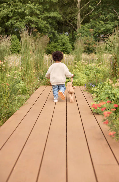 Kind loopt met biscuitkleurige Jollein Knuffel Bunny over houten pad in bloemenrijke buitenspeelomgeving.