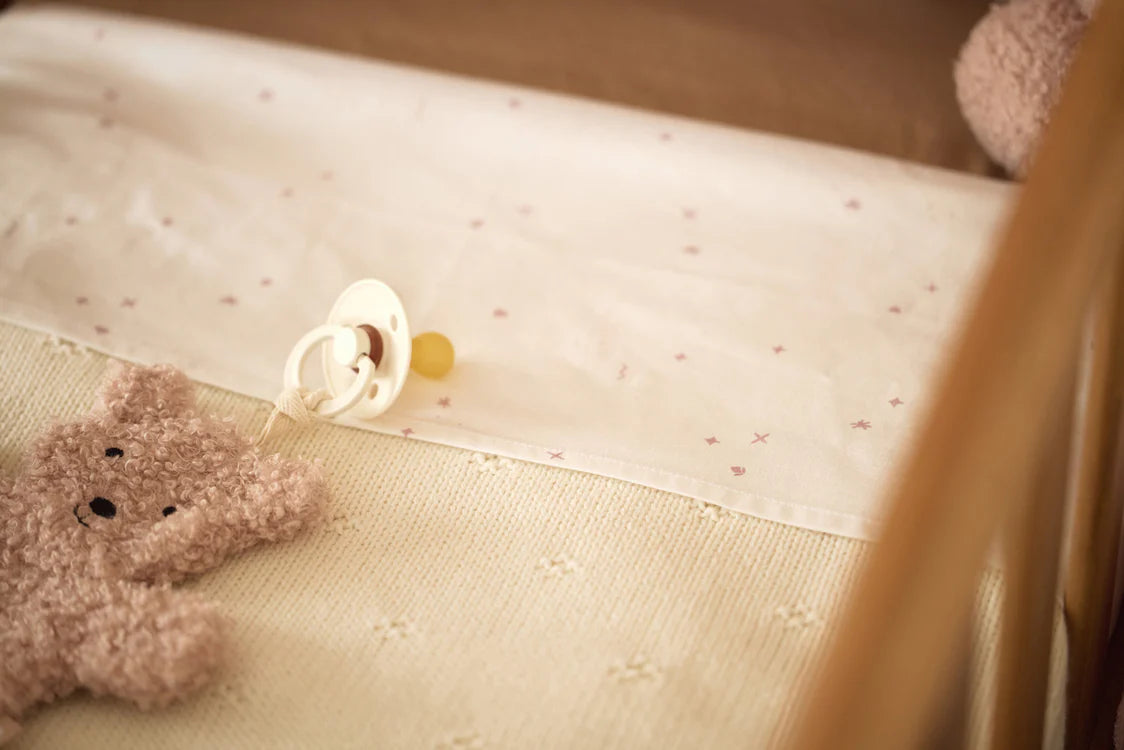 De Jollein Speenknuffel Teddy Bear Wild Rose ligt in een houten babyledikant met speen eraan.