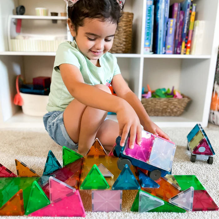 Een kind zit lachend op de grond te spelen met een kleurrijke Magnetic Magna Tiles Cars-set, terwijl de planken erachter vol staan met speelgoed.