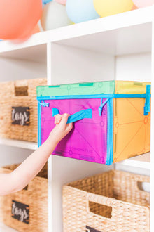 Een kind pakt een Magna Tiles opslagdoos op een witte plank, omringd door manden met de labels 'speelgoed' en magnetic tiles.