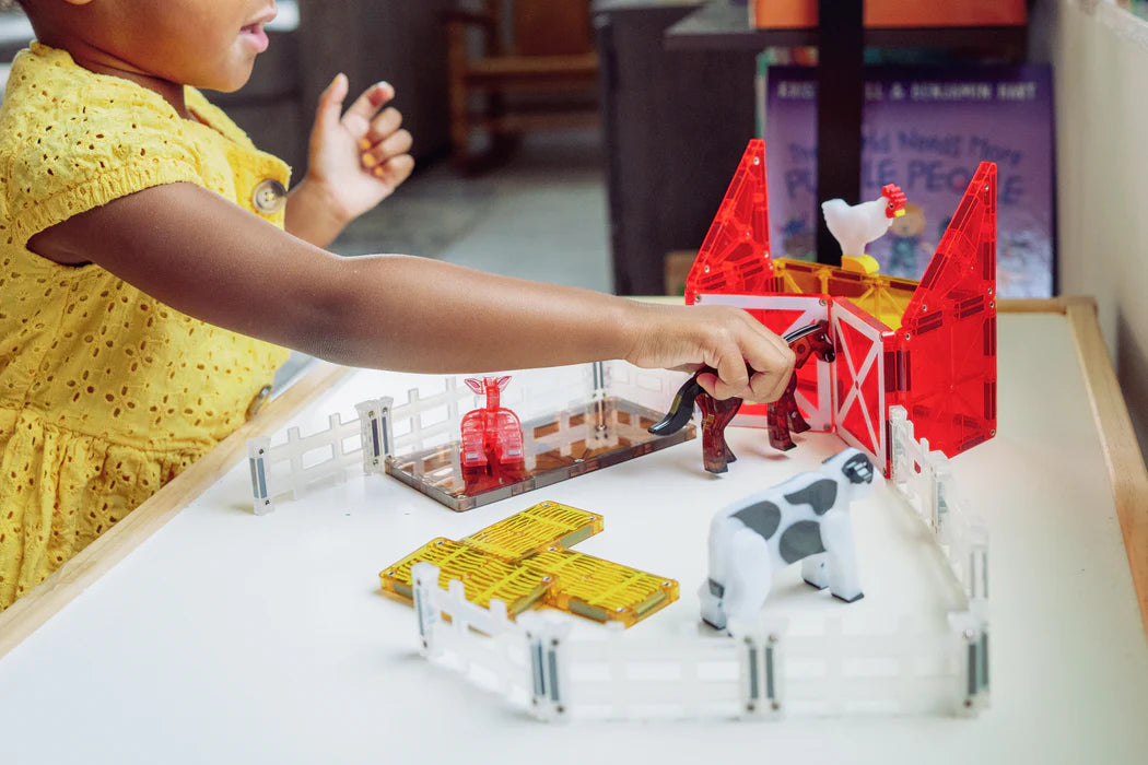 Een kind in een gele jurk speelt met de magnetic Magna Tiles boerderij dieren van magnetische bouwstenen op een tafel.