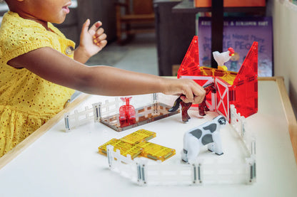 Een kind in een gele jurk speelt met de magnetic Magna Tiles boerderij dieren van magnetische bouwstenen op een tafel.