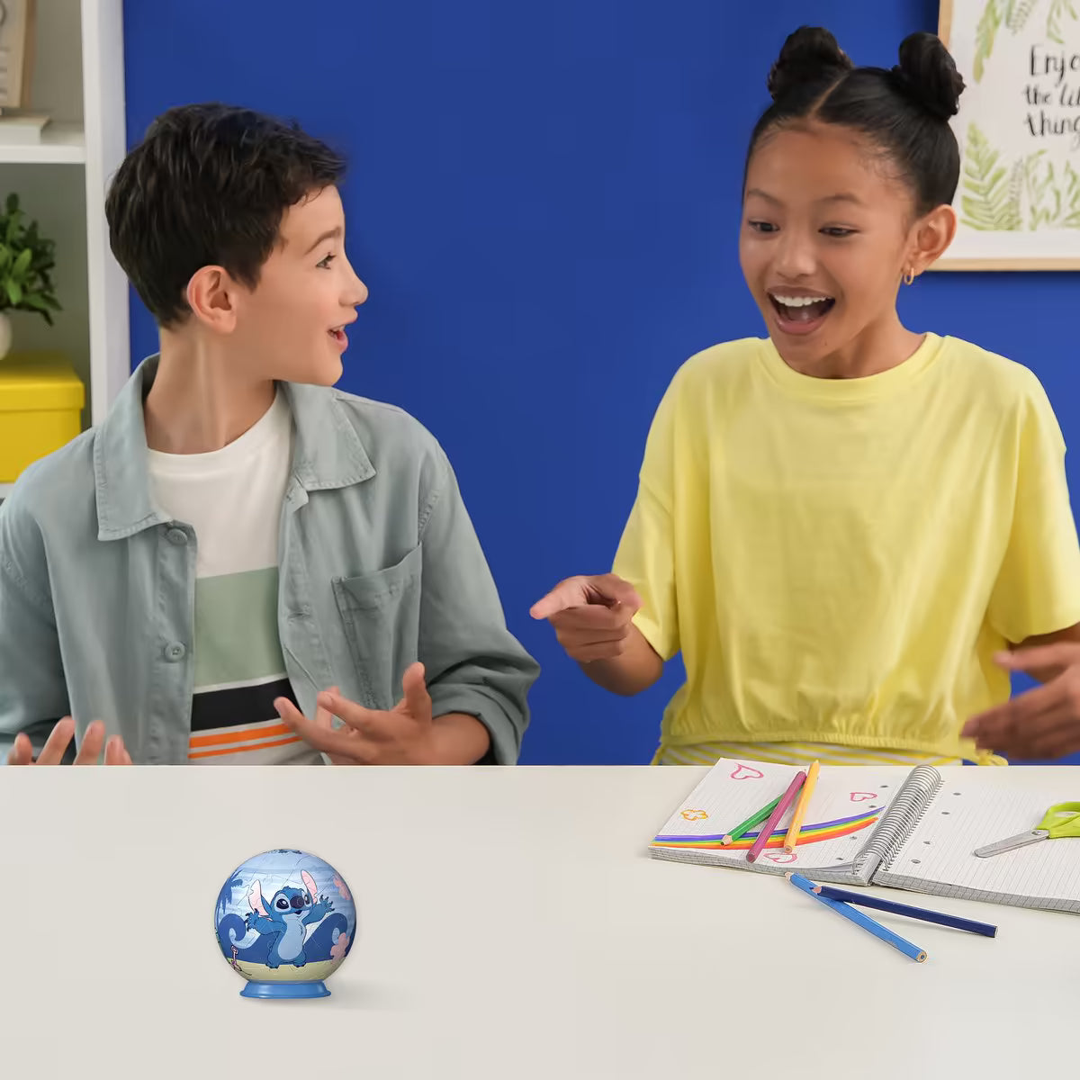 Twee kinderen lachen en wijzen enthousiast naar een afgeronde 3D Puzzle-Ball Disney Stitch op tafel, ideaal voor creatief speelplezier.