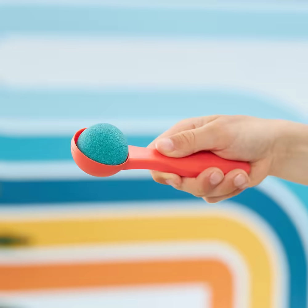 Hand met rode lepel en blauw Kinetic Sand bolletje tegen retro strepen, perfect sensorisch speelgoed voor kinderen.