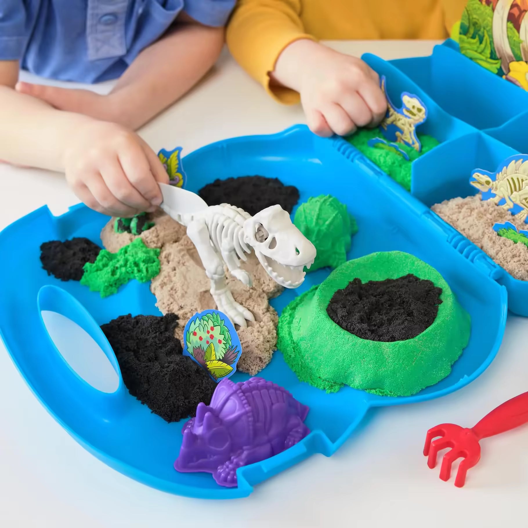 Close-up van de handen van kinderen die met de Kinetic Sand Trex Dino Case en dino-skeletten spelen, een voorbeeld van creatief speelgoed.