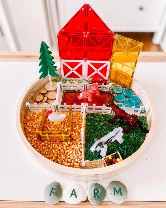 Een rond sensorisch dienblad met boerderijthema, met een speelgoedschuur, Magna Tiles-set, maïskorrels en stenen met de tekst 'FARM' erop.