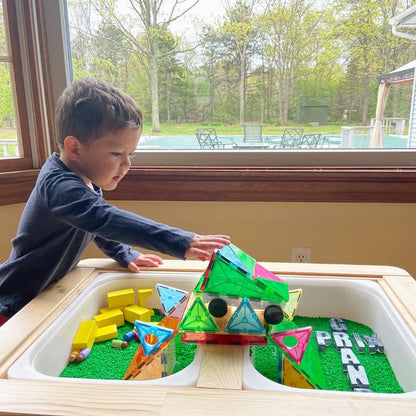 Een kind speelt met een kleurrijke Magnetic Magna Tiles Grand Prix-set bij een groot raam met uitzicht op een achtertuin met bomen.