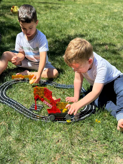 Twee kinderen spelen op het gras met een Magnetic Magna Tiles Builder-set. De een rangschikt de auto's terwijl hij de tegels verkent, de ander kijkt toe.
