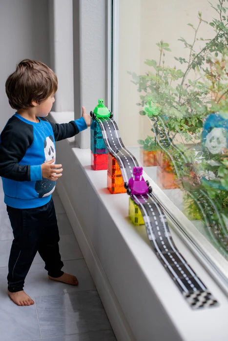 Een jongen speelt met Magnetic Magna Tiles Downhill Duo, met speelgoed autos op een zwarte racebaan voor het raam in kamer.