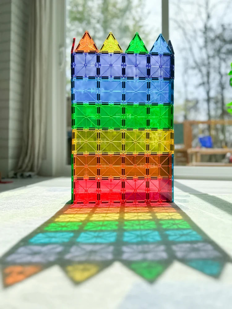 Hoge muur van magnetisch speelgoed van Magna Tiles in regenboogkleuren, verlicht door zonlicht met kleurrijke schaduwen op de vloer.