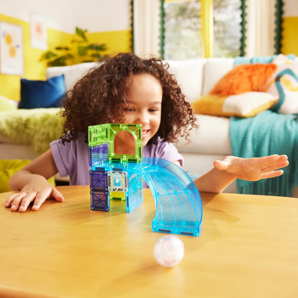 Kind rolt de Hamster Ball naar beneden over de blauwe glijbaan van de kleurrijke Magna Tiles speelset.