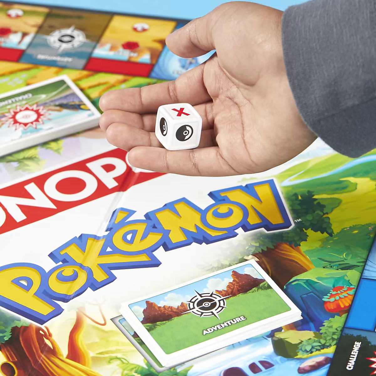 Detailfoto Monopoly Pokémon Hasbro bordspel met hand die witte dobbelsteen vasthoudt naast avontuurkaart op speelbord.